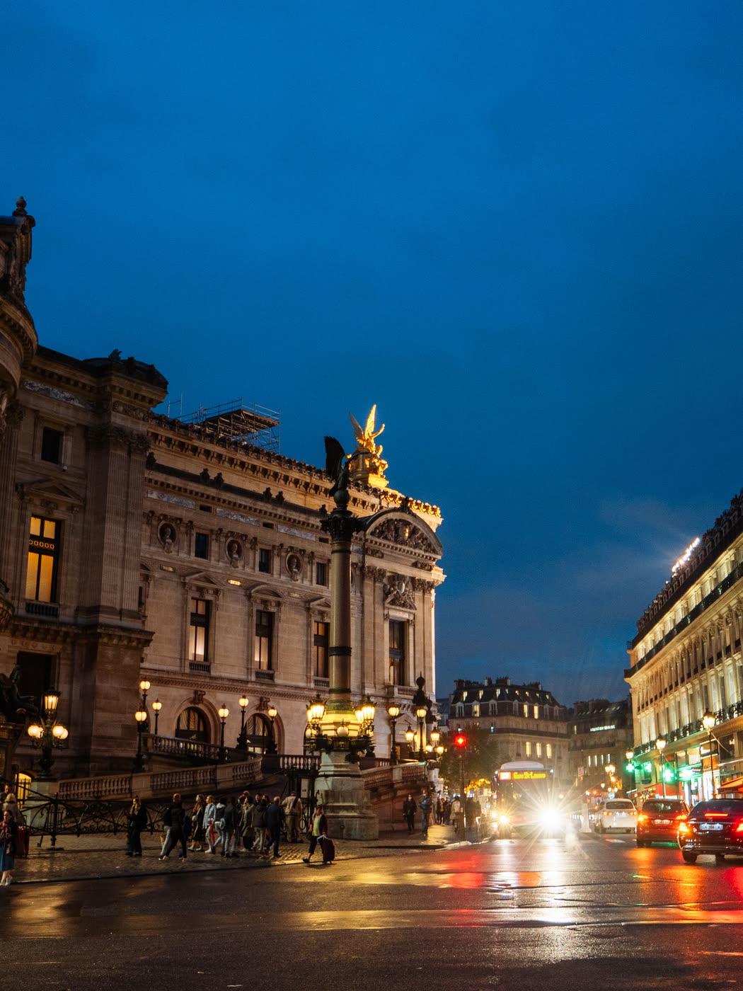 La façade de l’Opéra Garnier illuminée à l’heure bleue, et la circulation de la place de l’Opéra